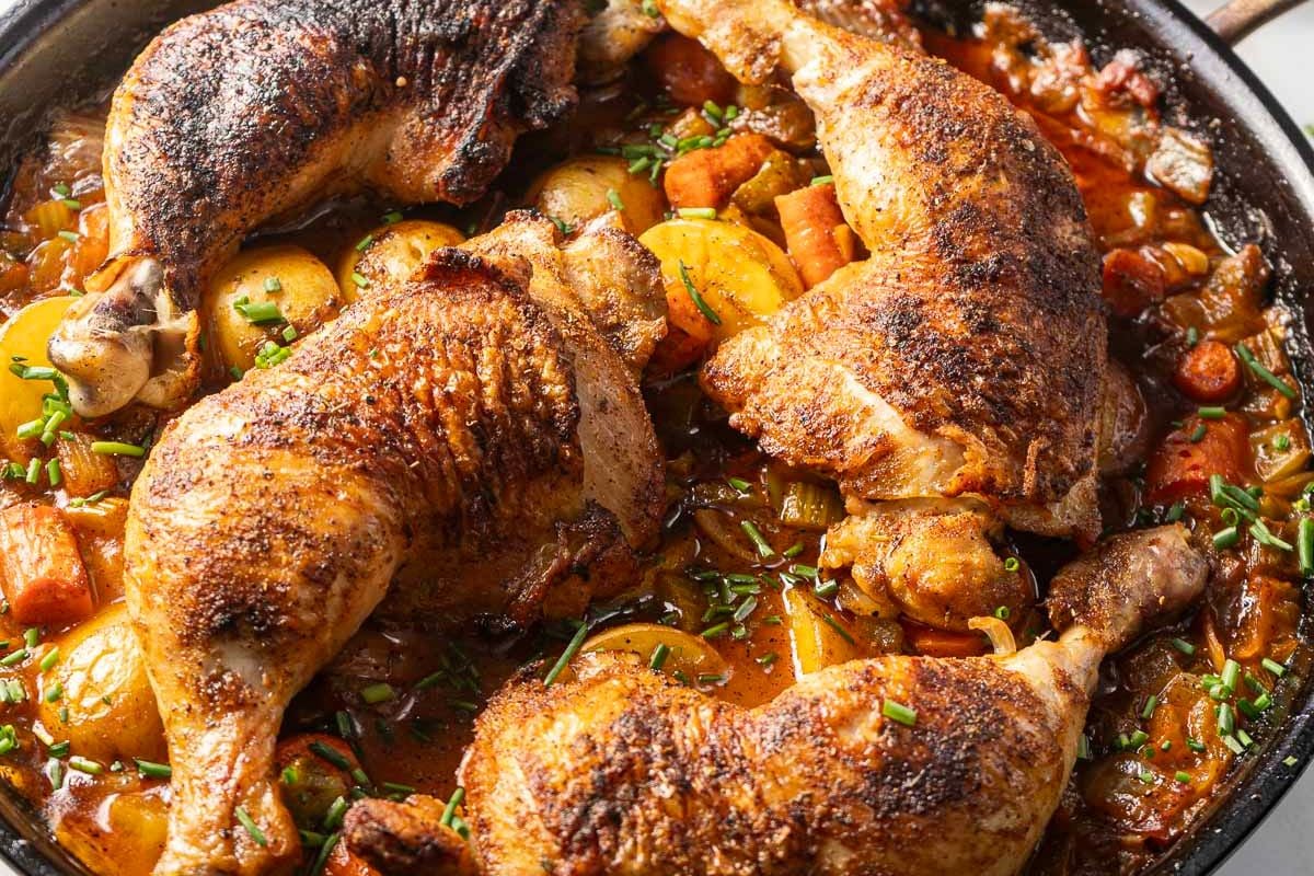 Four seasoned chicken legs are cooked in a skillet with potatoes, carrots, and a rich, tomato-based sauce, garnished with chopped herbs—making it one of the ultimate easy chicken recipes for weeknight meals.