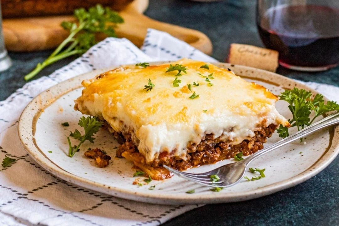 A piece of shepherd’s pie with melted cheese and parsley sits on a plate with a fork, next to a glass of red wine and a cork—a comforting reminder of classic recipes everyone should learn.