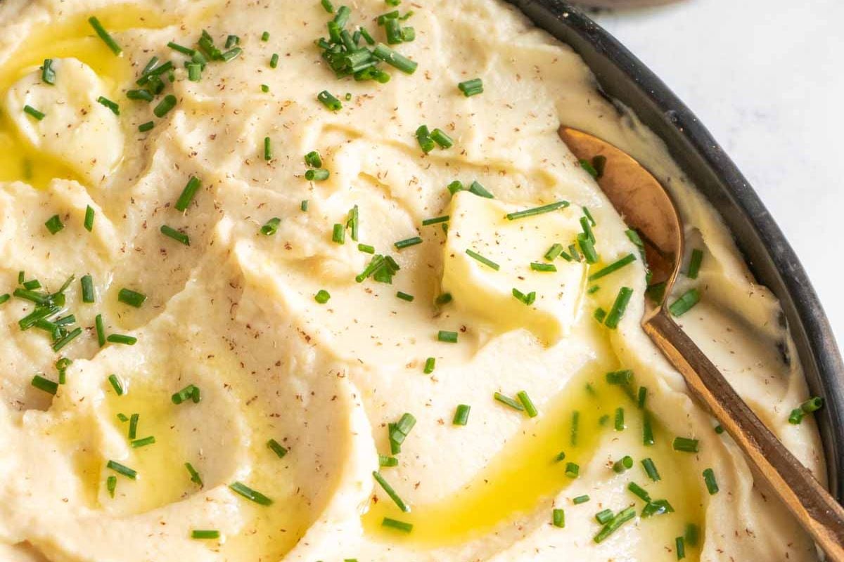 Bowl of mashed potatoes topped with melting butter, chopped chives, and black pepper, with a gold spoon resting in the dish—a classic favorite among Thanksgiving Side Dishes Recipes.