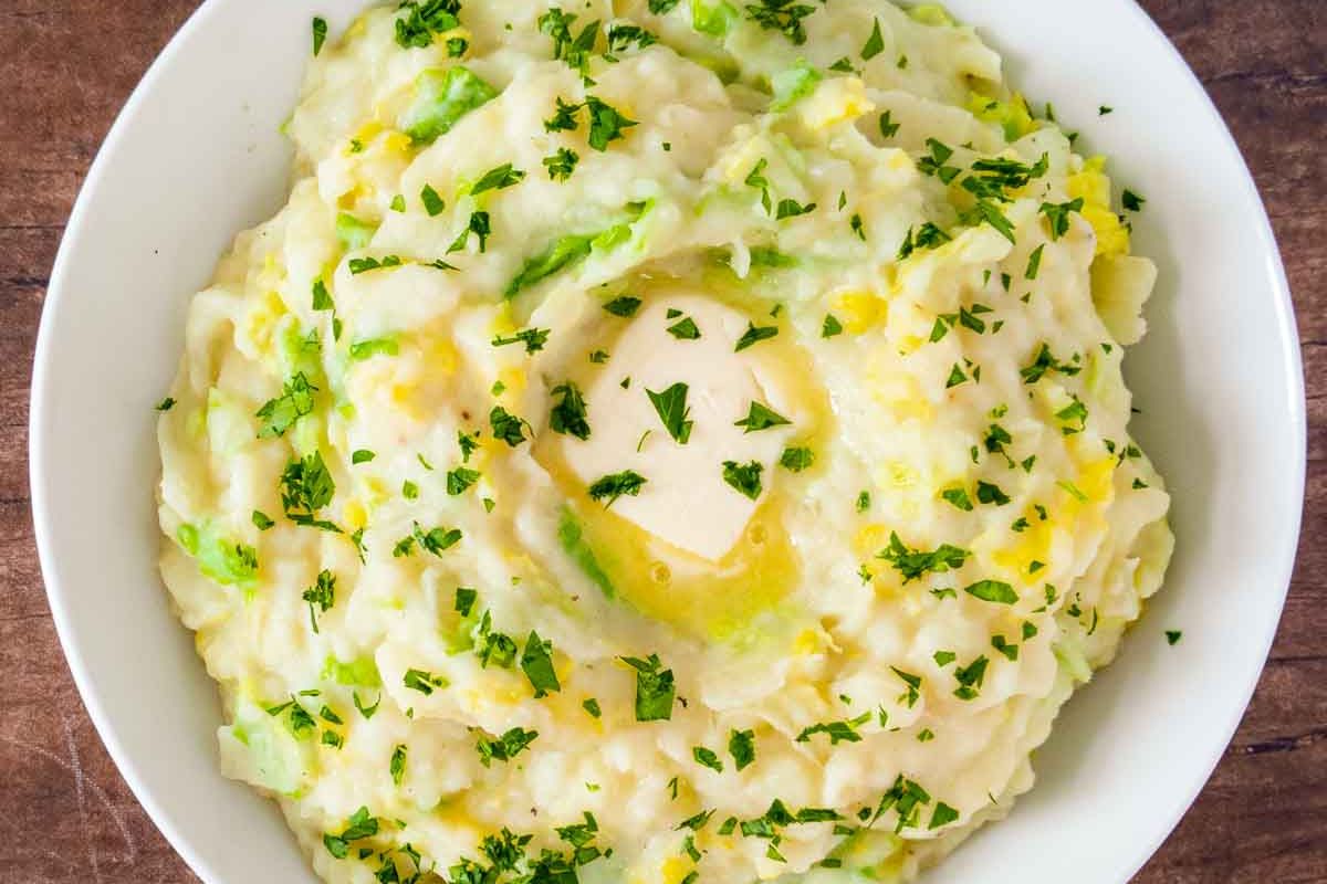 Bowl of mashed potatoes mixed with green cabbage and herbs, topped with a pat of butter and chopped parsley, on a wooden surface—a classic choice among comforting cabbage recipes.
