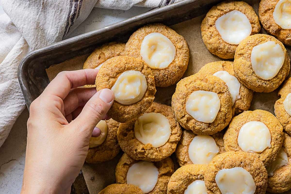 A hand holds a frosted cookie above a baking tray filled with gluten free pumpkin desserts on parchment paper.