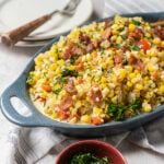 Casserole dish filled with creamed corn, chopped bacon, parsley, and diced vegetables, placed on a table with a stack of plates and a bowl of fresh herbs nearby.