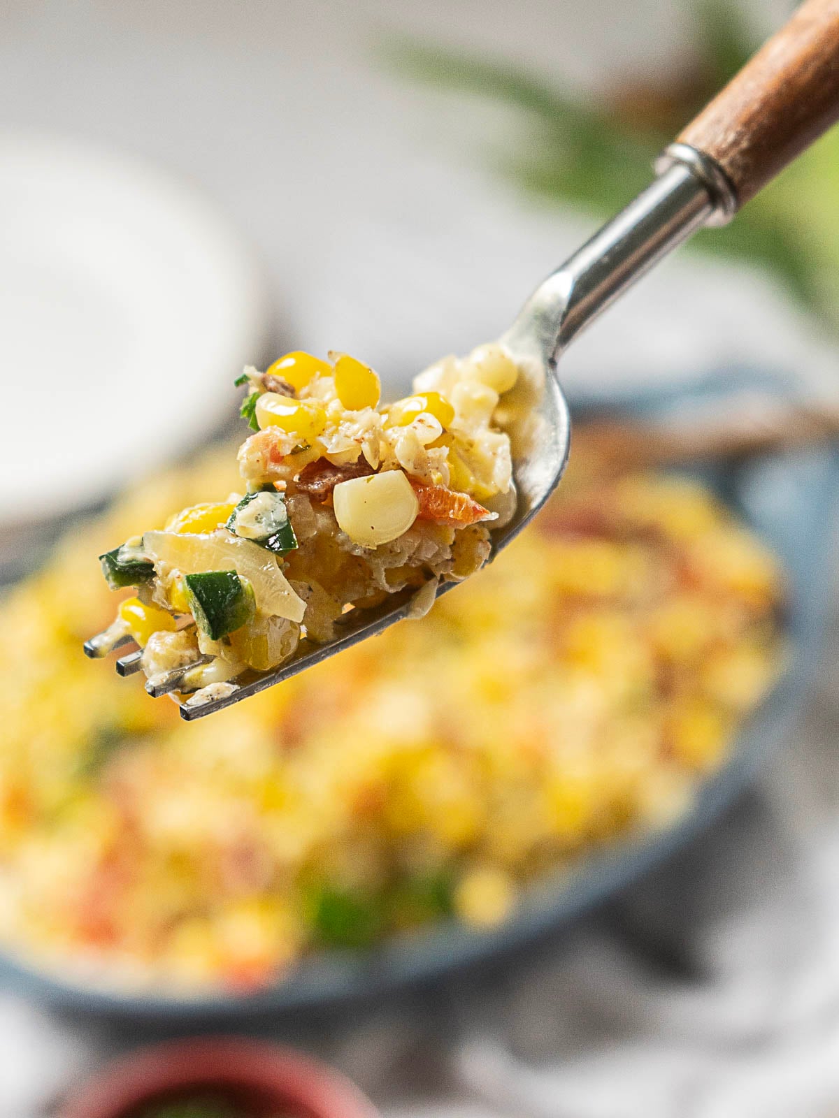 A close-up of a fork holding a bite of corn casserole with visible pieces of corn, green peppers, and other vegetables, with the rest of the dish blurred in the background.