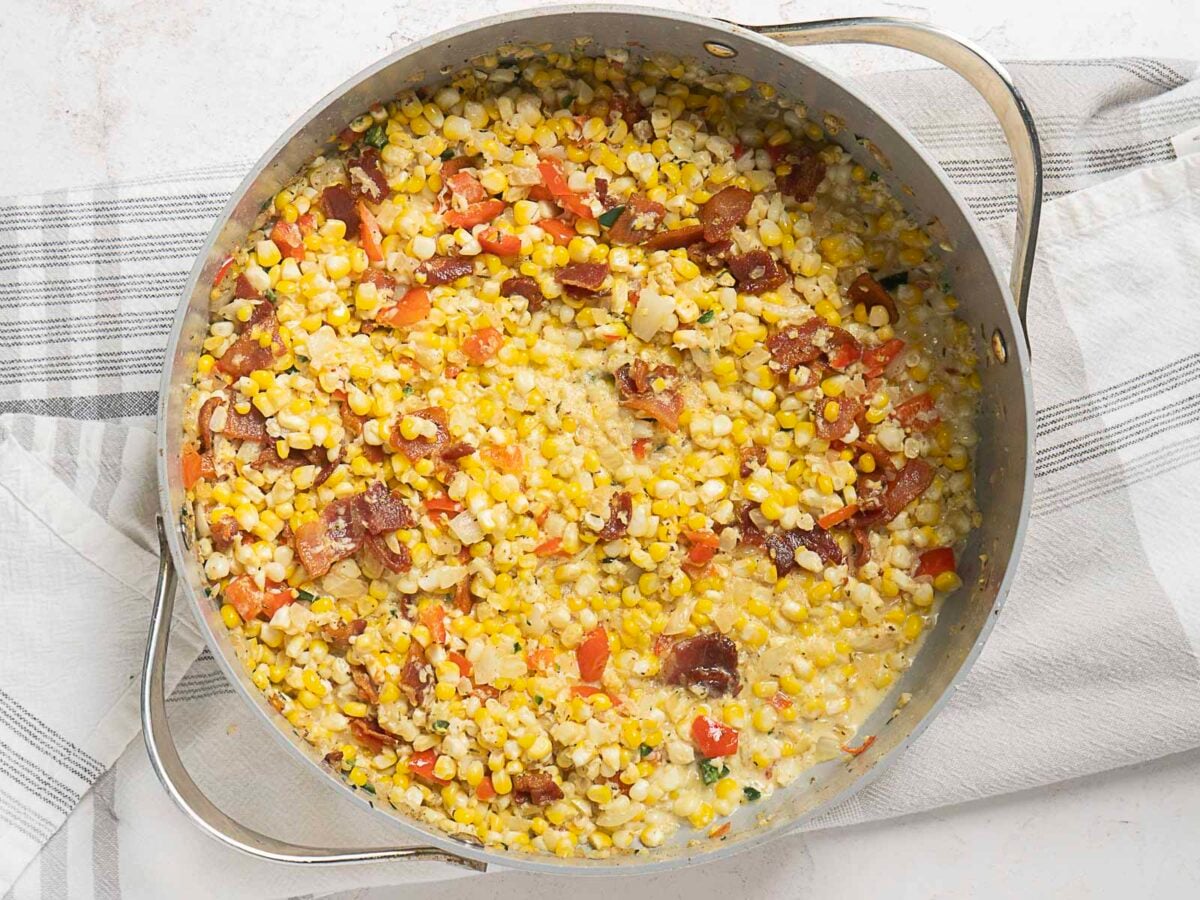 Large skillet filled with a mixture of corn, bacon, red bell peppers, and onions, resting on a striped kitchen towel.