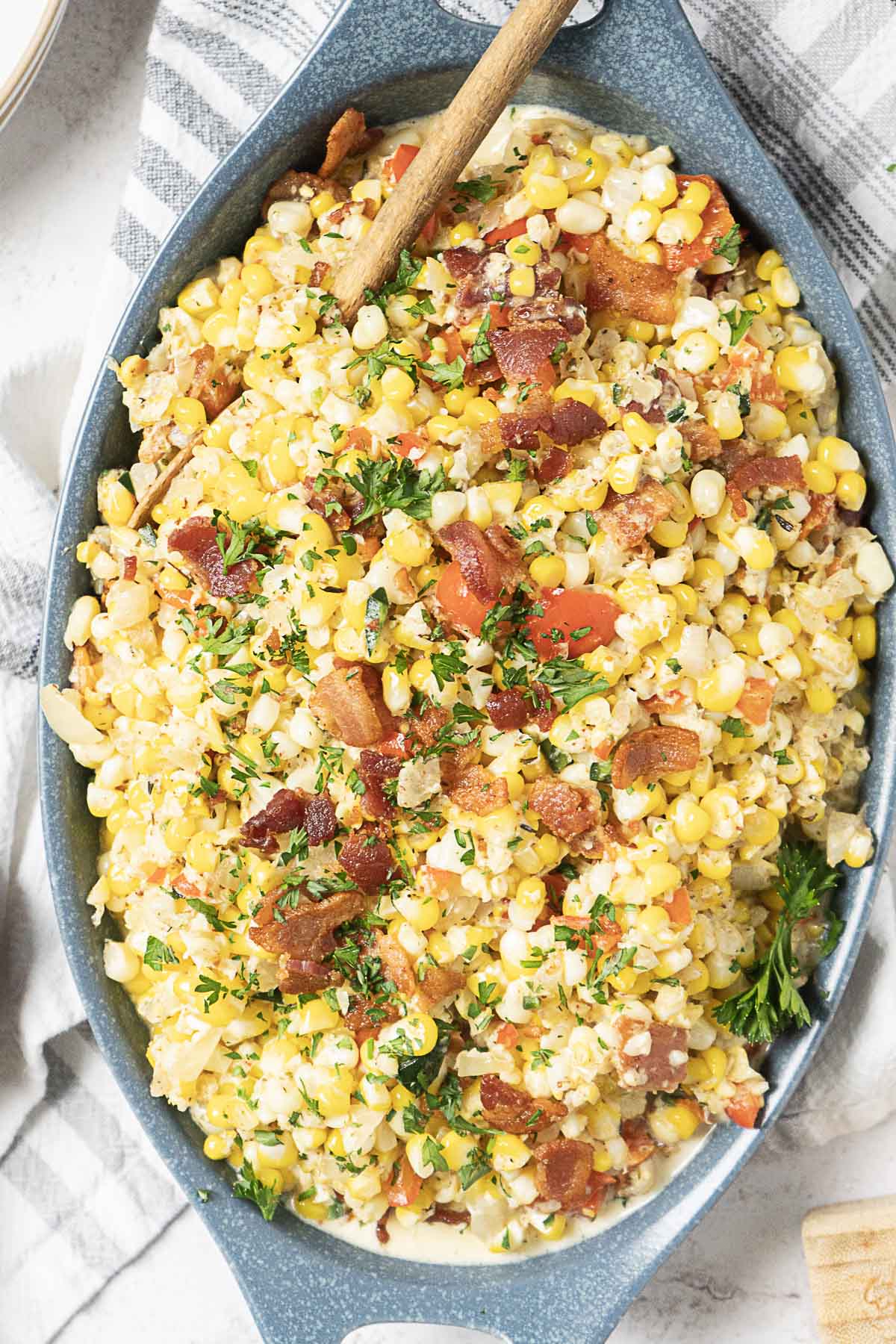 Oval dish filled with creamy corn maque choux mixed with chopped bacon, herbs, and diced vegetables, served with a wooden spoon.