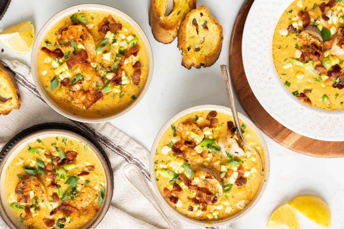 Four bowls of creamy soup, perfect for fall recipes, are garnished with shrimp, bacon, corn, and herbs, served alongside toasted bread slices and lemon wedges on a light surface.