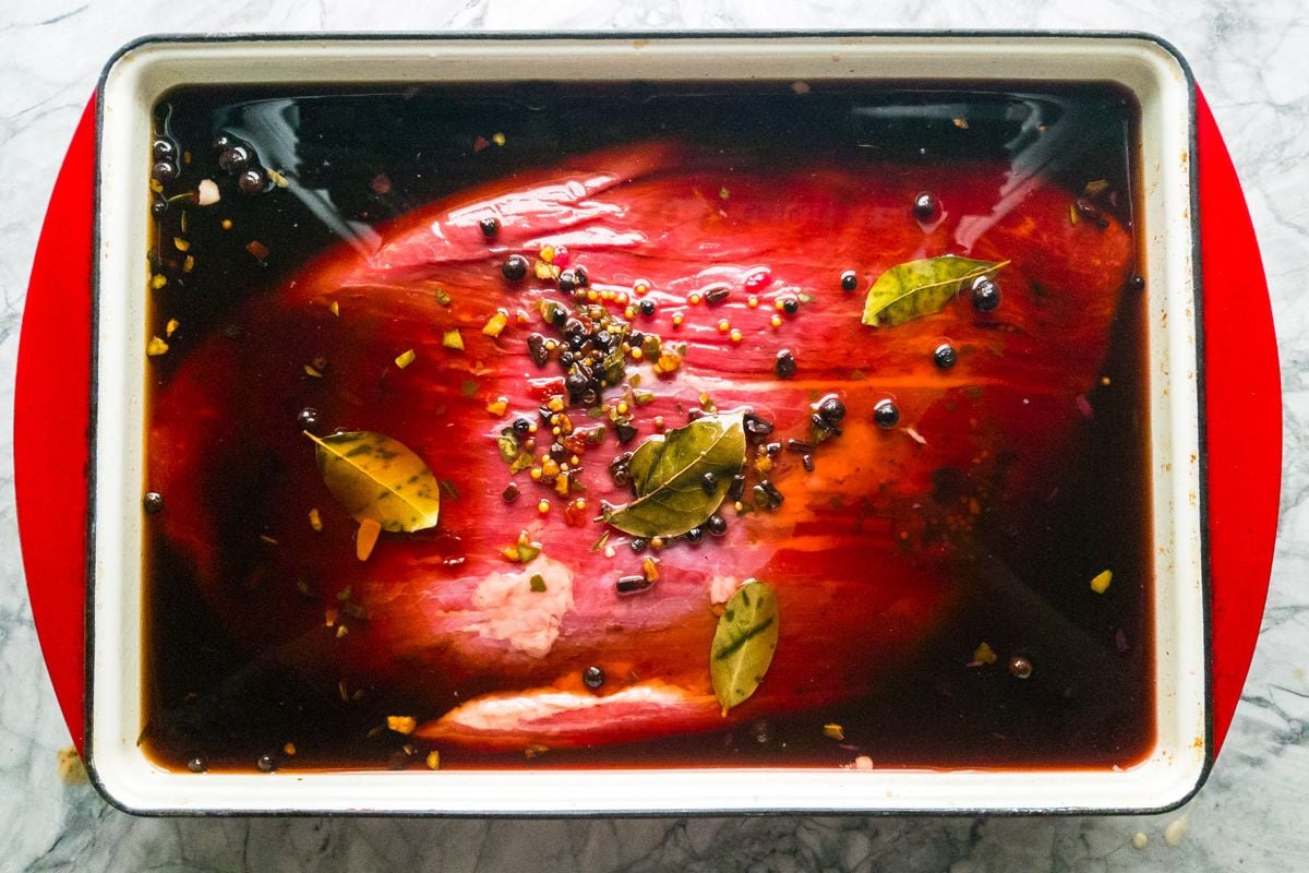 A large cut of raw beef is marinating in a rectangular dish with dark liquid, whole spices, peppercorns, bay leaves, and chopped garlic on top—perfect for easy recipes that make delicious family meals.