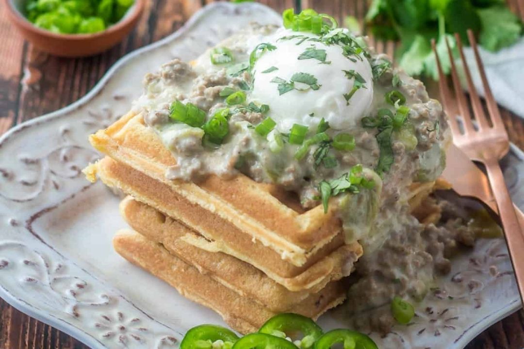 Stack of waffles topped with creamy sausage gravy, a dollop of sour cream, chopped green onions, and fresh herbs, served on a decorative plate with jalapeño slices on the side—perfect for anyone seeking savory breakfasts or creative breakfast ideas.