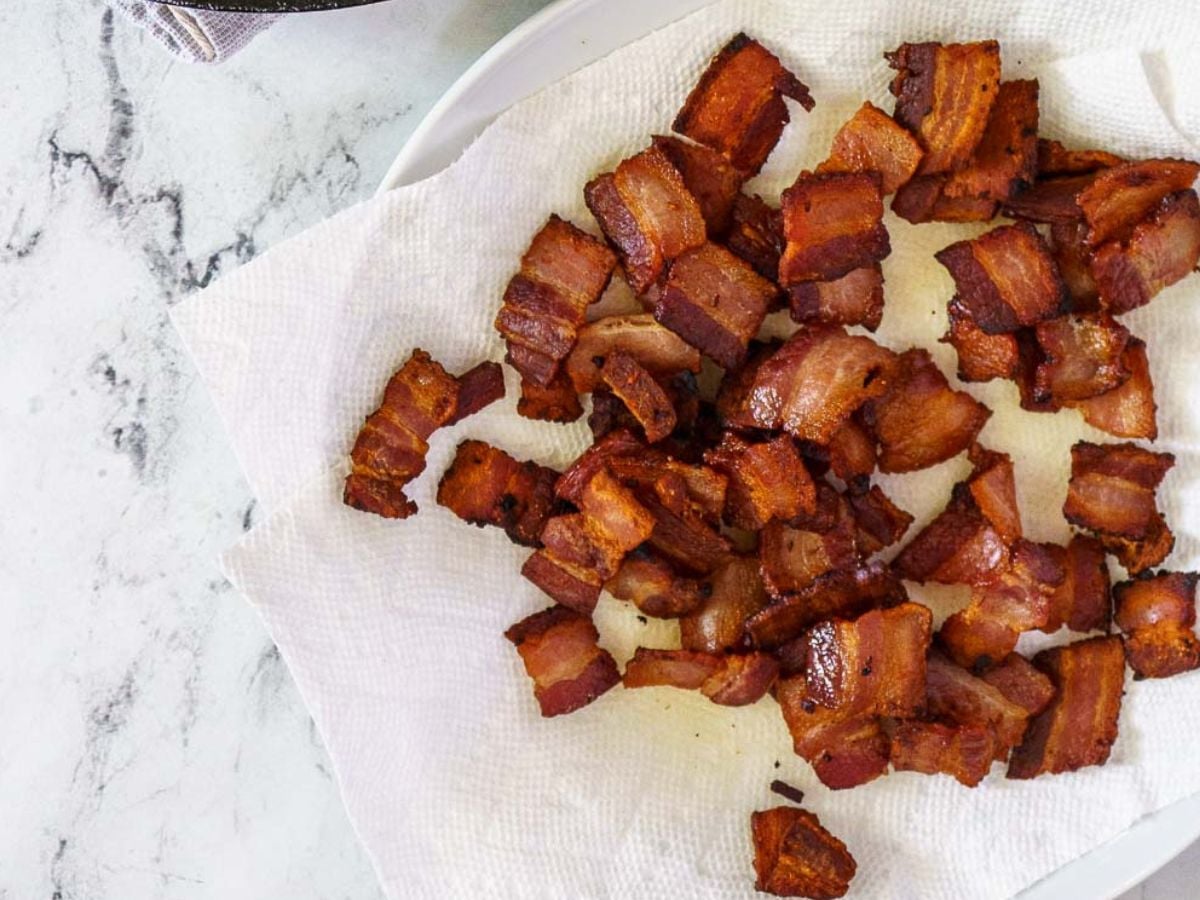 Crispy cooked bacon pieces are spread on a white paper towel atop a plate, with a marble countertop in the background.
