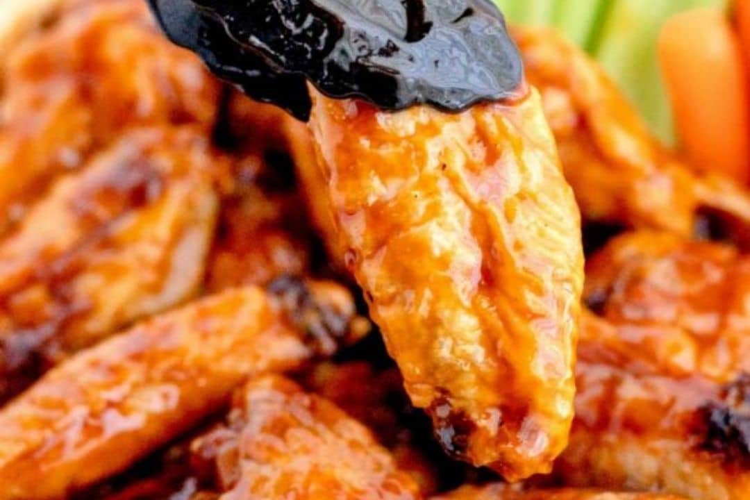 Close-up of saucy chicken wings, with one wing being dipped into a dark sauce—perfect halftime snacks. Carrot and celery sticks are visible in the background.