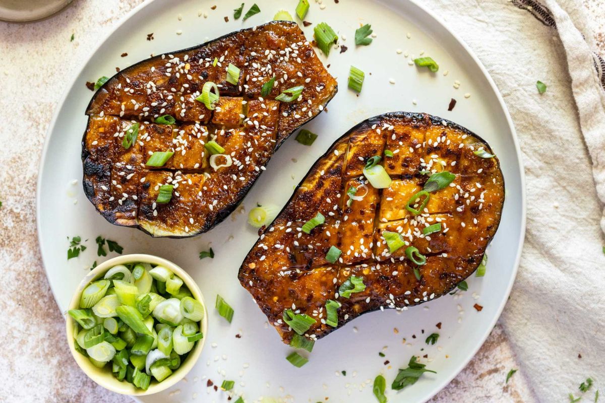 Two roasted eggplant halves, flavor-packed and garnished with sesame seeds and chopped green onions, are served on a plate alongside a small bowl of sliced green onions—perfect for adding to your collection of standout side dishes.