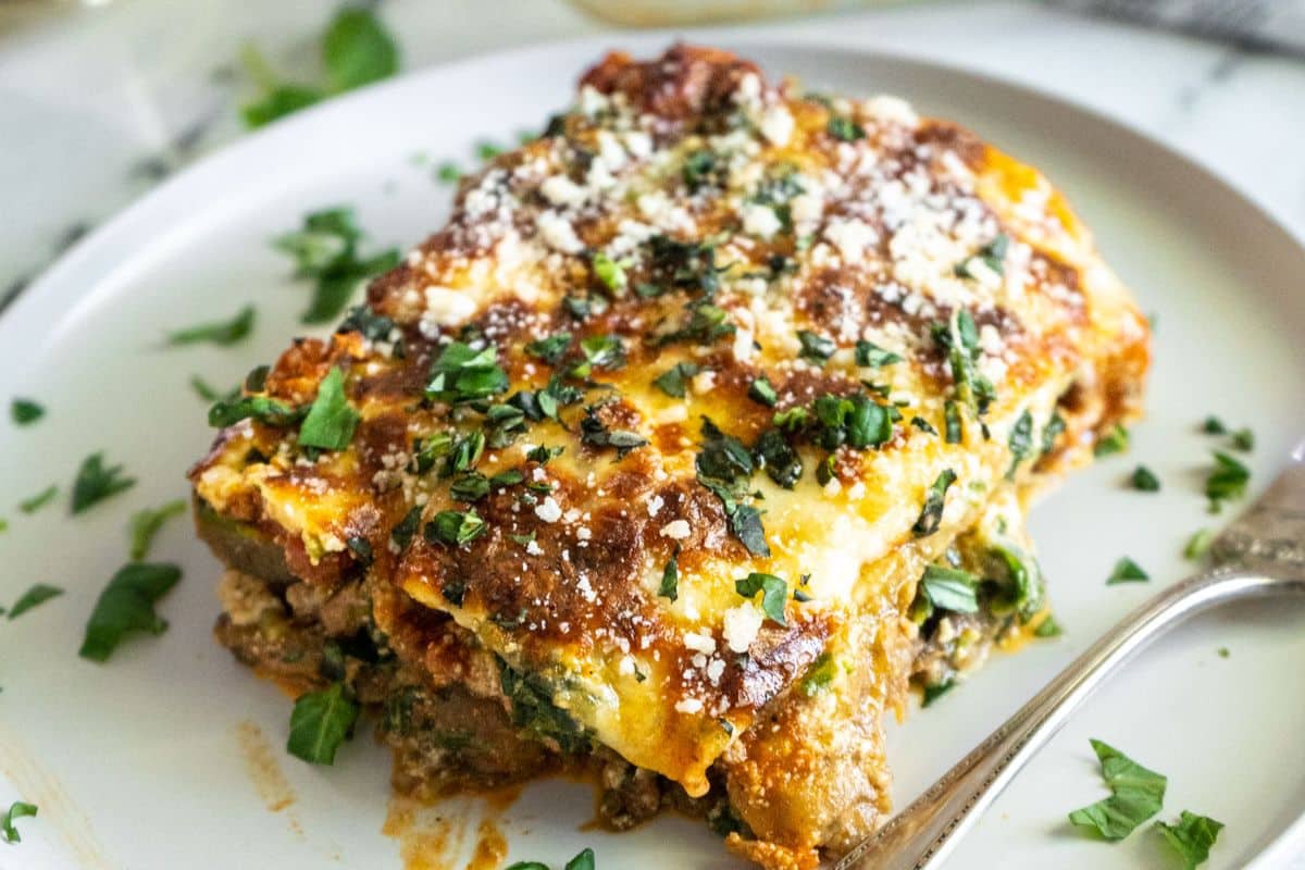 A slice of baked lasagna, perfect for family meals, is topped with chopped parsley and grated cheese on a white plate with a fork.