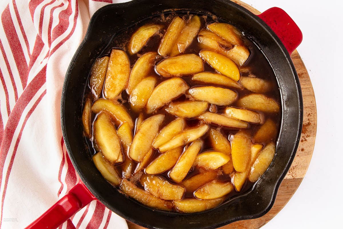 Sliced apples cooked in a dark syrup inside a red-handled skillet, placed on a wooden board next to a red and white striped towel—a beautiful addition to your Thanksgiving table and perfect for anyone seeking delicious side dish recipes.