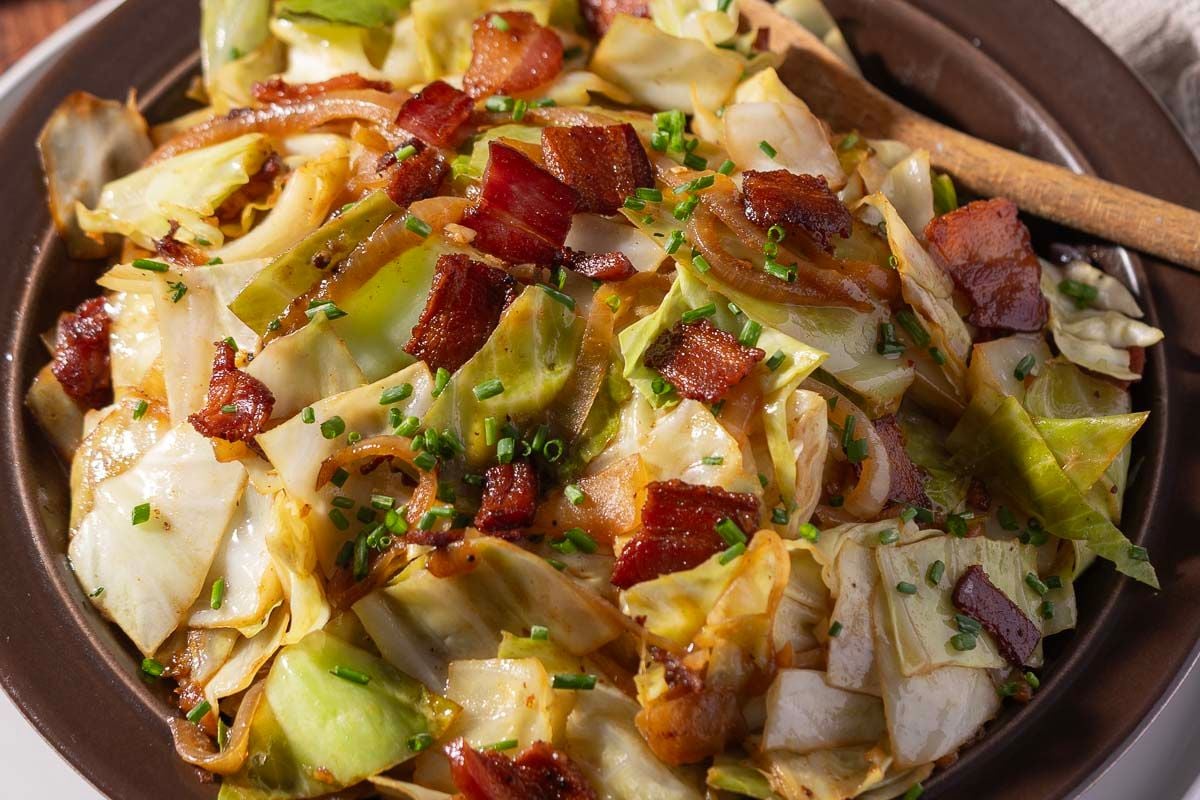 A plate of cooked cabbage mixed with crispy bacon pieces, garnished with chopped chives, showcases one of the simplest yet tastiest cabbage recipes. A wooden spoon rests on the side.