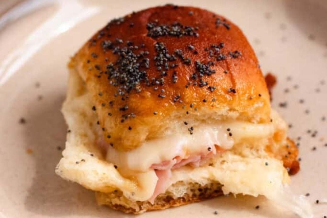 A close-up of a ham and cheese slider with melted cheese, ham, and poppy seeds on a soft, golden brown bun, served on a beige plate—one of those stress free potluck recipes that’s perfect to feed a crowd.