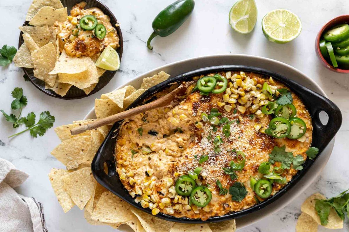 Oval dish of cheesy corn dip topped with jalapeno slices and cilantro, surrounded by tortilla chips; lime halves and jalapeno slices on the side—perfect for potluck recipes and sure to feed a crowd with stress free preparation.