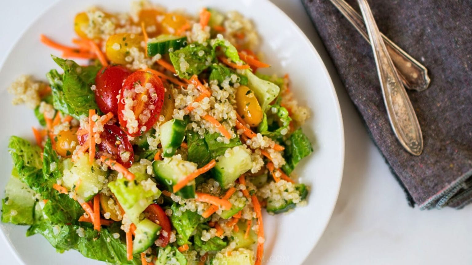 Plate of quinoa salad with lettuce, cucumbers, cherry tomatoes, carrot strips, and avocado—perfect for salad season. Two forks rest on a dark napkin beside the plate.