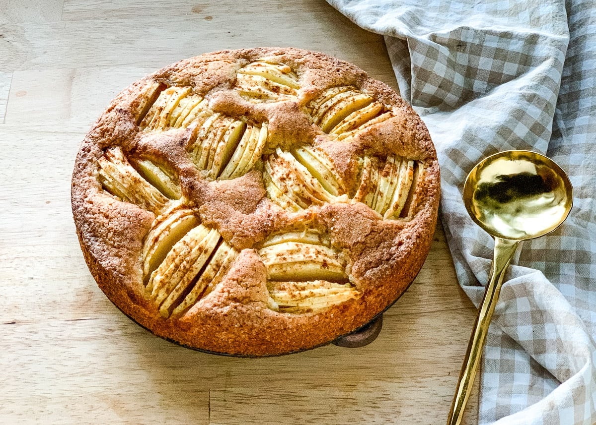 A round apple cake—one of the classic German favorites—with visible apple slices on top, sits next to a gold spoon and a checkered cloth on a light wooden surface.