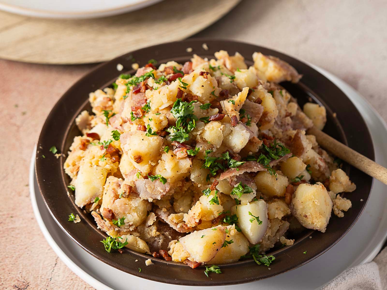 A dark plate of German potato salad with chopped bacon, parsley, and a light dressing—a true German favorite—served with a wooden spoon on a white plate. This classic example of German food is perfect for celebrating Oktoberfest or any hearty meal.