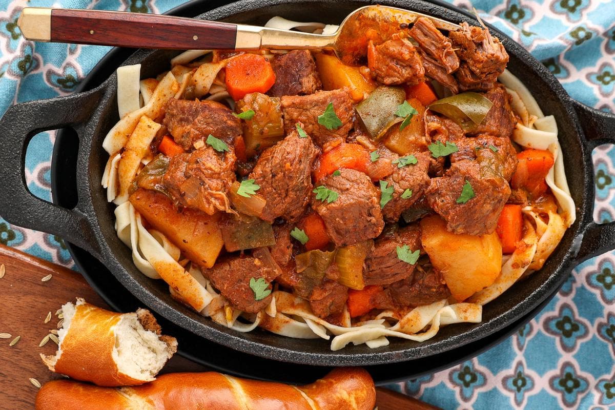 A black dish filled with beef stew, potatoes, carrots, and green peppers—one of those hearty German favorites—served over wide noodles and garnished with parsley, with a bread roll and patterned cloth nearby. This comforting meal is perfect for celebrating Oktoberfest or exploring classic German recipes at home.