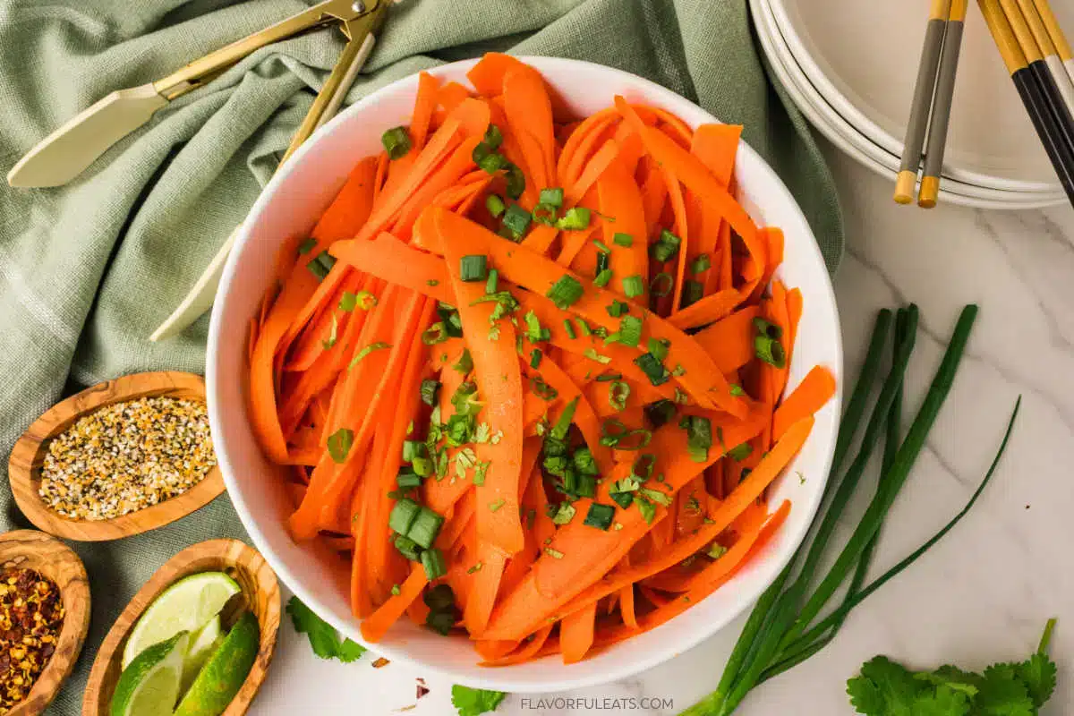 A white bowl filled with sliced carrots topped with green onions, perfect for easy carrot recipes, surrounded by seasoning bowls, lime wedges, chopsticks, and a green cloth.