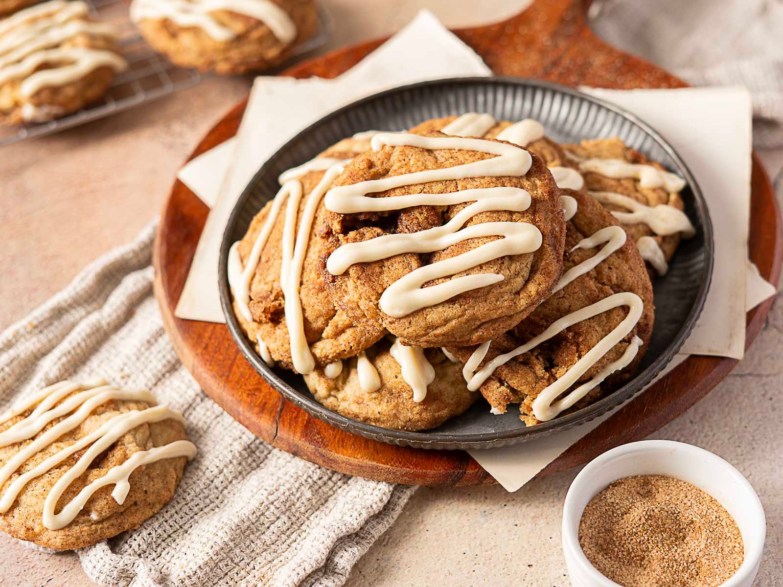 A plate of cookies with white icing drizzle sits on napkins, surrounded by the cozy flavors of fall recipes—a small bowl of cinnamon sugar and more gluten free cookies fill the background, making this a perfect treat for a gluten free fall.