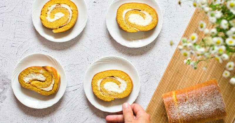 Four plates with gluten free pumpkin roll cake slices on a white surface, one slice being picked up, with a full cake roll and flowers on the side.