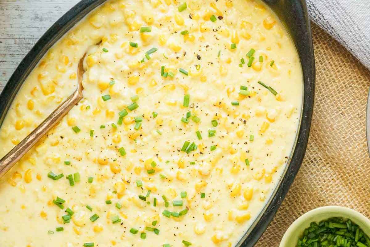 A dish of creamy corn casserole, one of those timeless recipes, topped with chopped chives and black pepper, with a wooden spoon resting in the casserole.