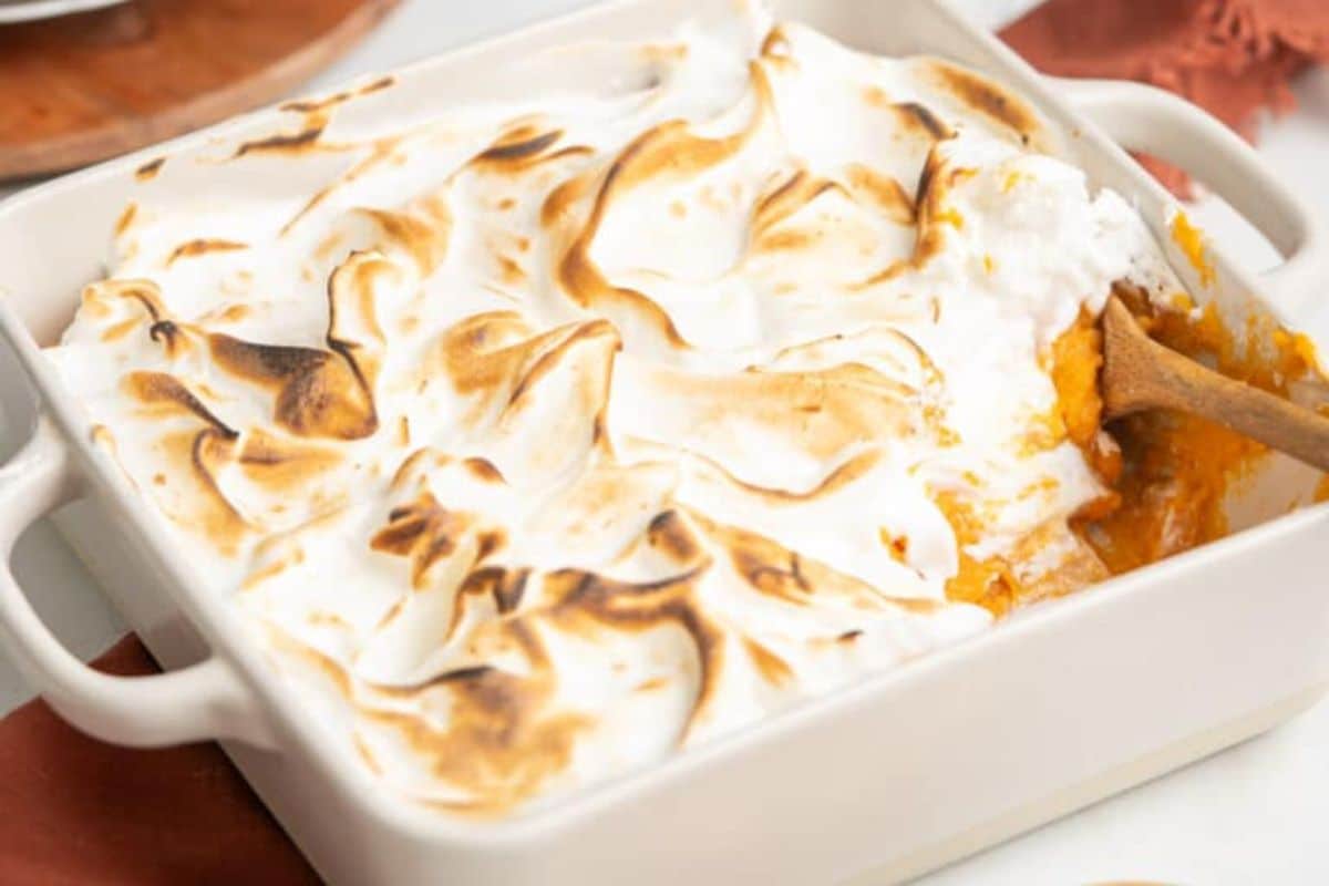 A square baking dish filled with sweet potato casserole topped with toasted meringue, with a wooden spoon scooping out a portion—perfect for anyone searching for unique casserole recipes to save for later.