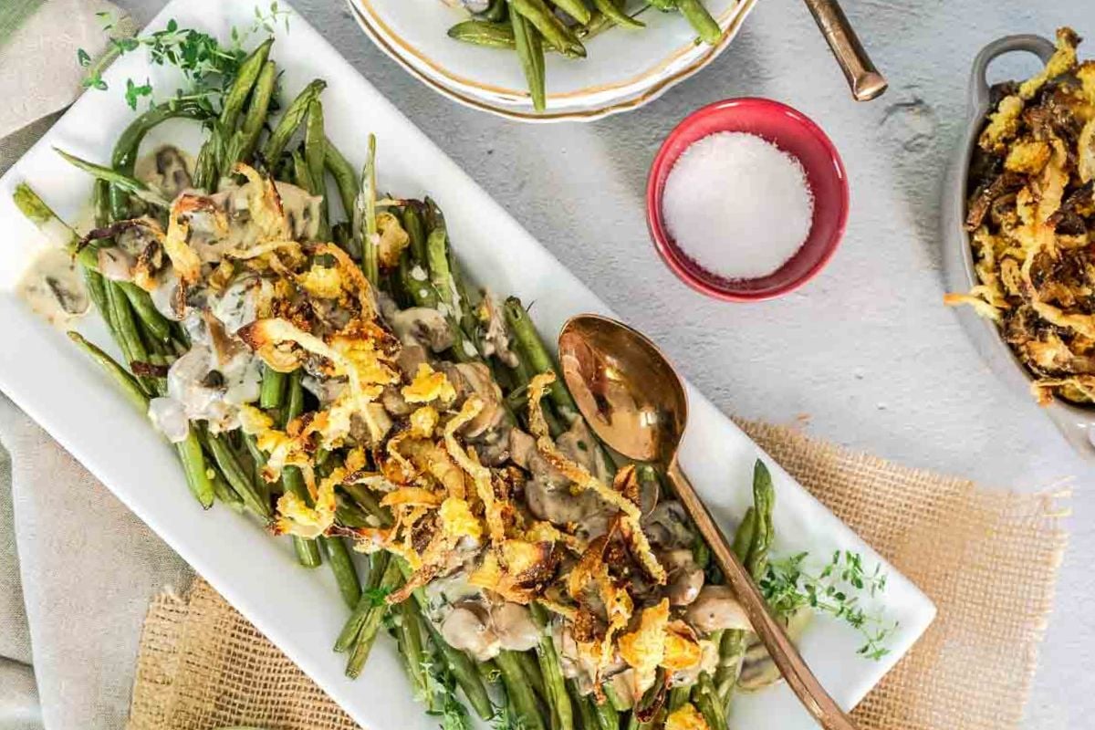 Green beans topped with creamy mushroom sauce and crispy fried onions are served on a rectangular white platter with a gold spoon, surrounded by flavor-packed side dishes and a small bowl of salt—an irresistible recipe for any gathering.