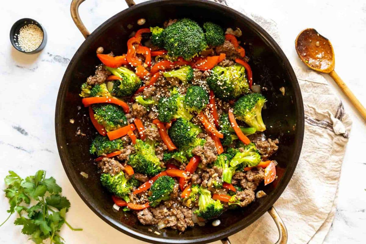 A skillet filled with cooked ground beef, broccoli florets, and red bell peppers, topped with sesame seeds—a vibrant addition to your collection of Dinner Recipes. A wooden spoon, cilantro, and extra sesame seeds are nearby.