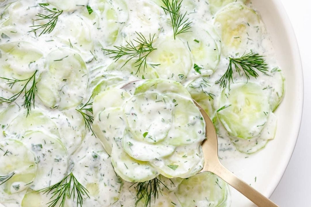 Sliced cucumber salad mixed with creamy dressing and fresh dill, served in a white dish with a spoon—one of the classic German Favorites, perfect for adding freshness to your Oktoberfest Recipes.