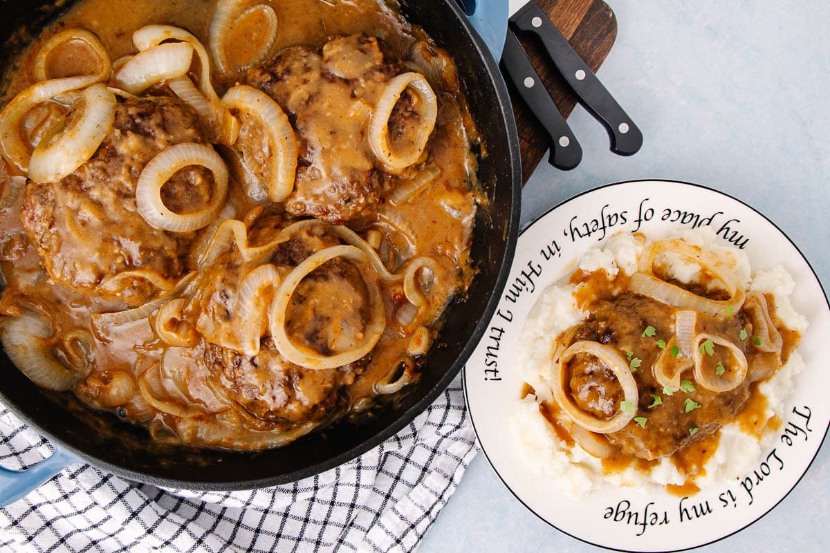 A skillet with Salisbury steak and onions in brown gravy, inspired by classic Southern recipes, sits next to a plate with mashed potatoes, gravy, and steak, garnished with herbs.