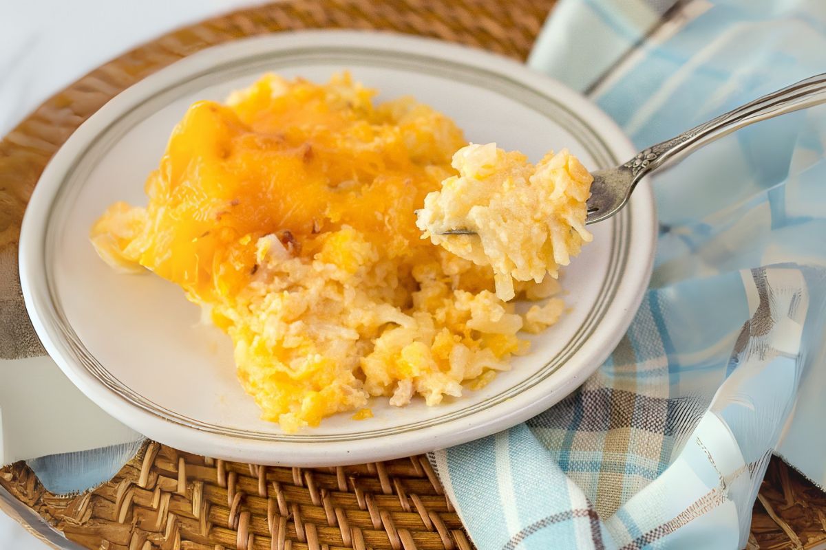 A serving of cheesy hash brown casserole on a white plate, with a fork lifting a bite—an irresistible addition to your Thanksgiving spread. The plate is on a woven mat next to a blue and white plaid cloth, making it one of the most comforting side dishes for any holiday table.