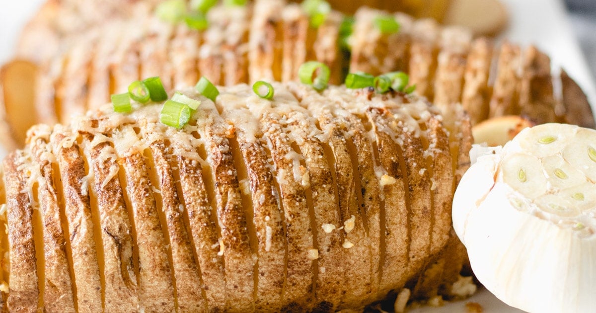 Sliced baked potatoes topped with melted cheese and chopped green onions make one of the ultimate potato side dishes, served with a roasted garlic bulb on the side—perfect for cozy fall dinner gatherings.