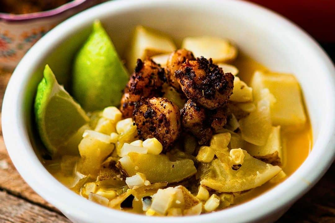 A white bowl filled with chopped vegetables, grilled seasoned shrimp, lime wedges, and corn sits on a wooden surface—perfect inspiration for autumn recipes or cozy soup recipes to warm up chilly evenings.