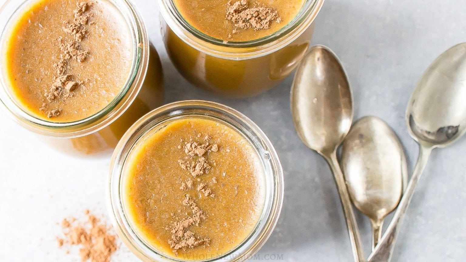 Three glass jars filled with a brown pumpkin dessert, each topped with cocoa powder, sit beside three metal spoons and a small pile of cocoa powder on a light surface.