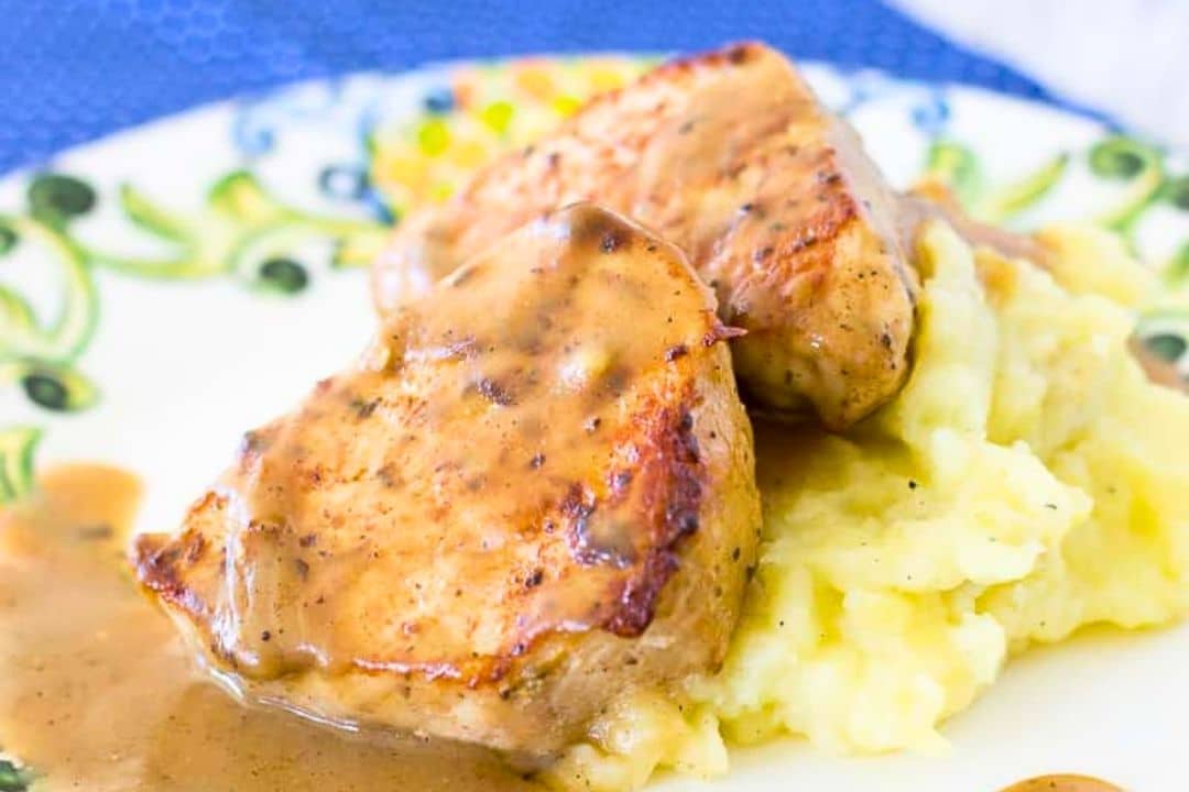 Two pieces of grilled pork with brown gravy served on mashed potatoes, presented on a decorative plate—a classic comfort food perfect for Sunday suppers and those who crave home cooked meals.