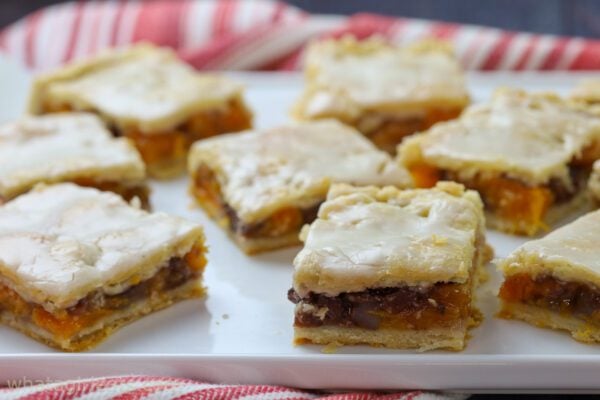 Nine glazed square bars with a layered filling of fruit and nuts are arranged on a white plate with a red-striped towel underneath—an ideal stress-free option to feed a crowd at your next gathering or potluck.