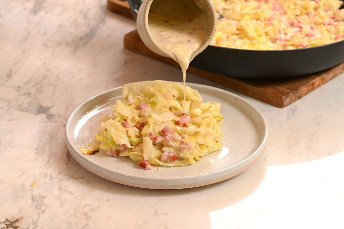 A serving of creamy cabbage and ham on a plate, with extra sauce being poured over it from a small pitcher—a delicious addition to your collection of comforting cabbage dishes.