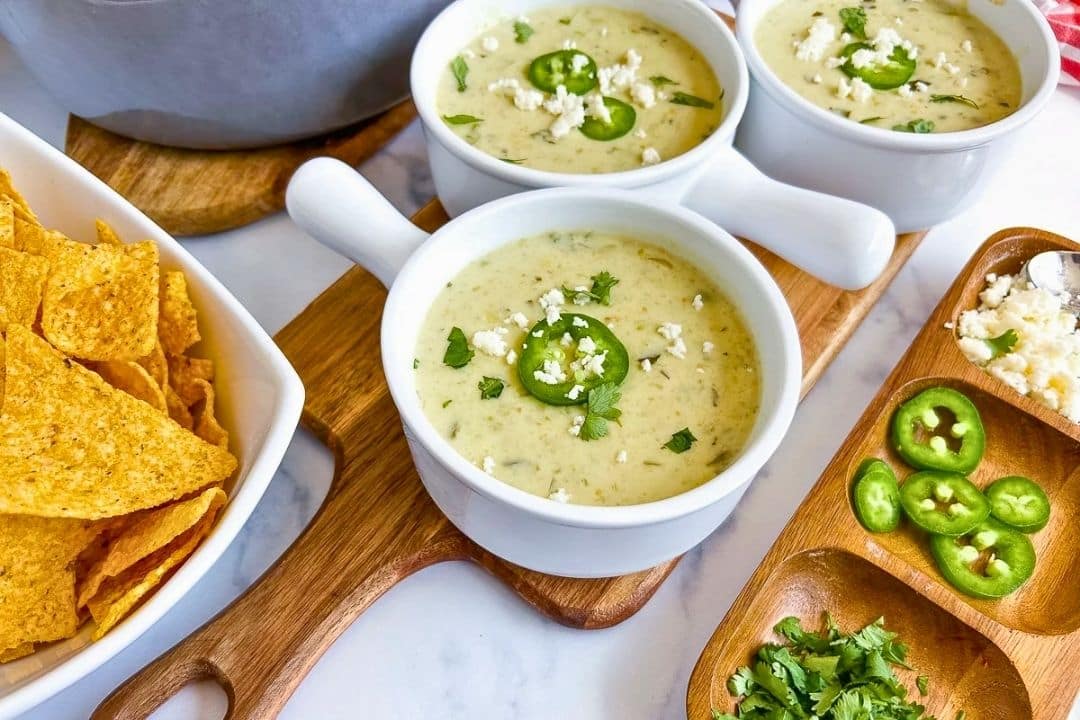 Three white bowls of creamy soup, perfect for cool nights, are garnished with jalapeño, cheese, and cilantro atop wooden boards. Beside them sits a dish of tortilla chips and small trays filled with sliced jalapeños, cheese, and cilantro—an inviting scene ideal for your favorite fall recipes or cozy soup recipes.