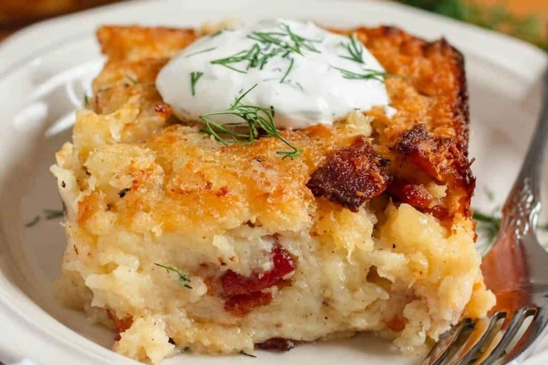 A slice of cheesy potato casserole with bacon, topped with sour cream and fresh dill, served on a white plate with a fork—this cozy dish is perfect for fall dinners and a must-try for anyone who loves hearty potato side dishes.