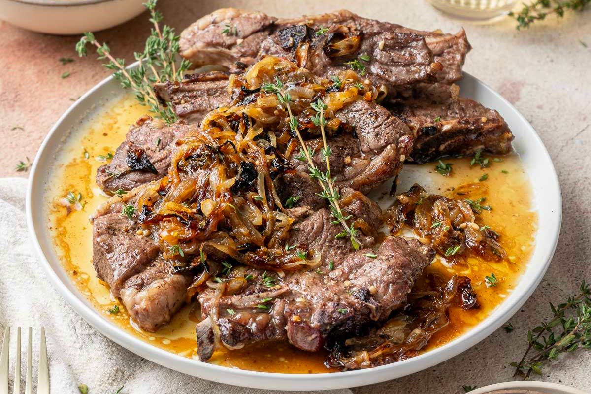 Plate of cooked steak topped with caramelized onions and fresh herbs, served with sauce on a white dish—perfect for those seeking popular dinner ideas or looking to add something delicious to their saved recipes.