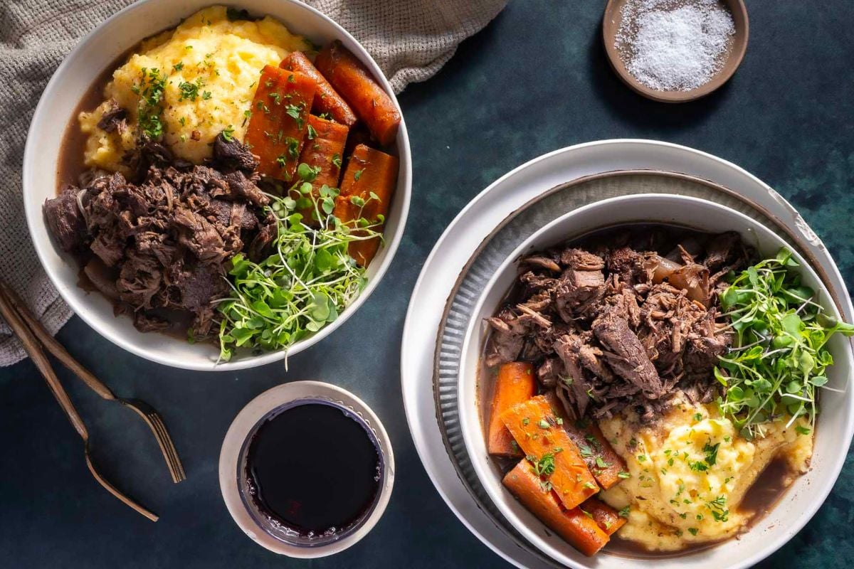 Two bowls of shredded beef with mashed potatoes, carrots, and fresh herbs—a perfect Cozy Braises meal for Sunday Nights, served with red wine and gold cutlery.