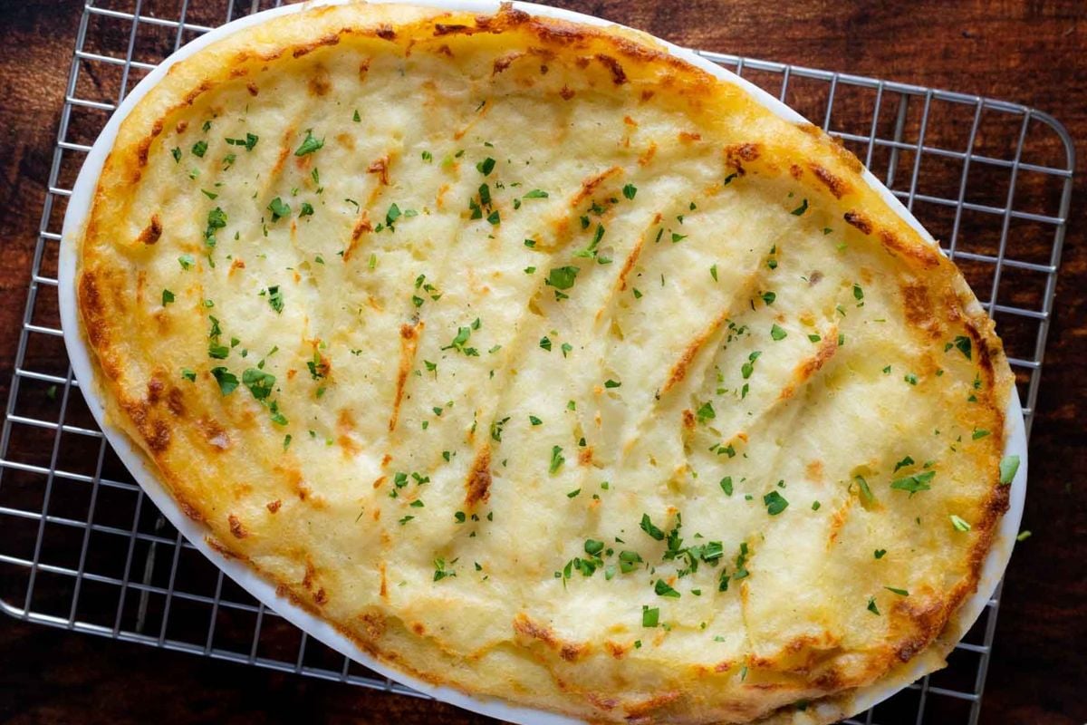 An oval dish of baked shepherd’s pie topped with golden mashed potatoes and sprinkled with chopped herbs, cooling on a wire rack—a comforting favorite among classic recipes and a must-try for anyone looking for essential recipes to learn.
