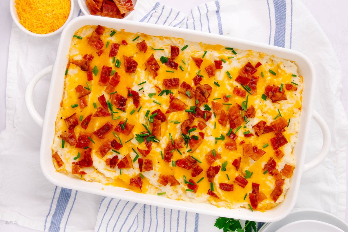A white baking dish filled with mashed potatoes topped with melted cheese, bacon, and chives—perfect for weeknight meals or easy make ahead recipes.
