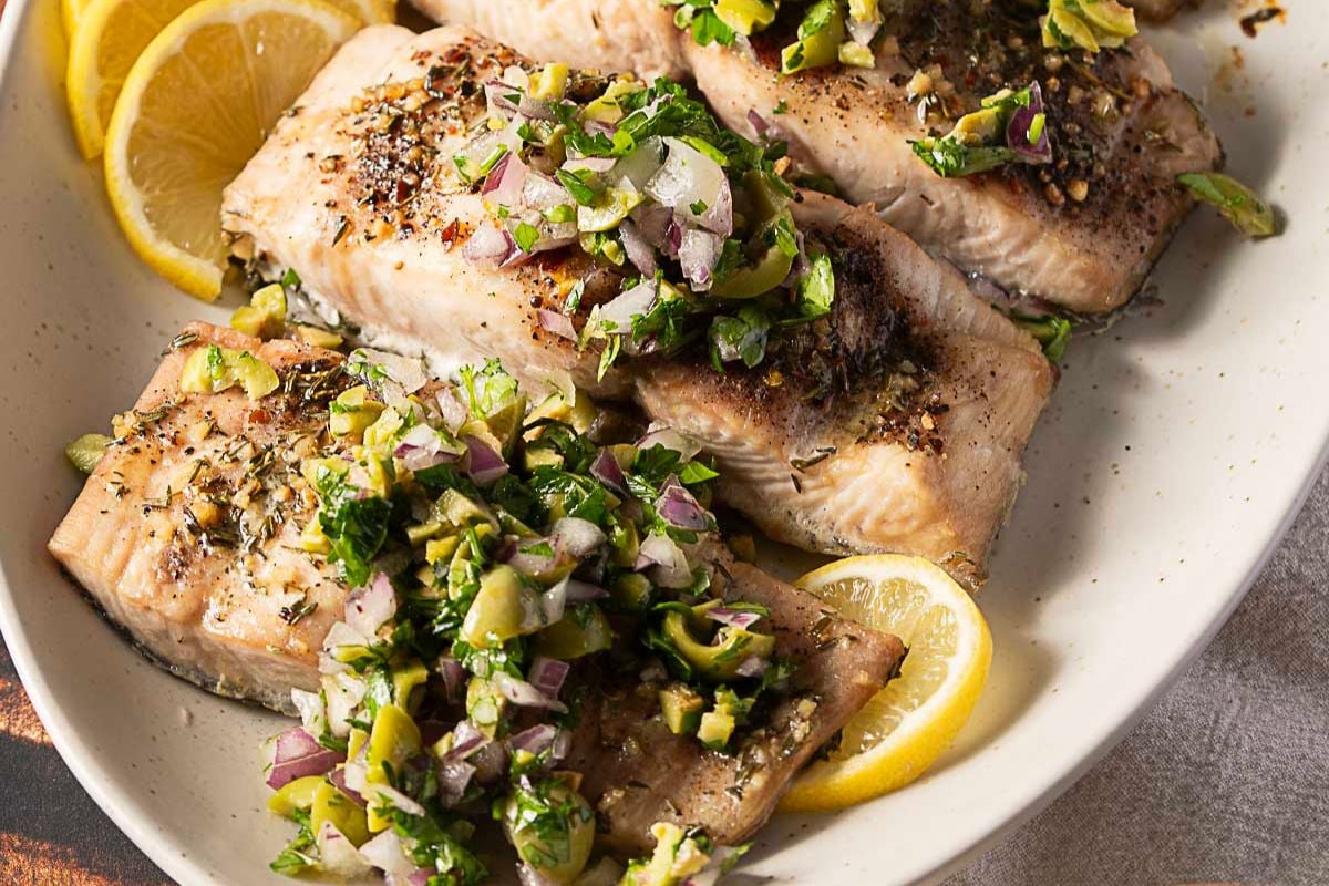 Three pieces of baked salmon fillet topped with a chopped olive, onion, and herb relish, garnished with lemon slices on a white oval plate—perfect for those seeking easy fish dishes or looking to expand their collection of delicious seafood recipes.