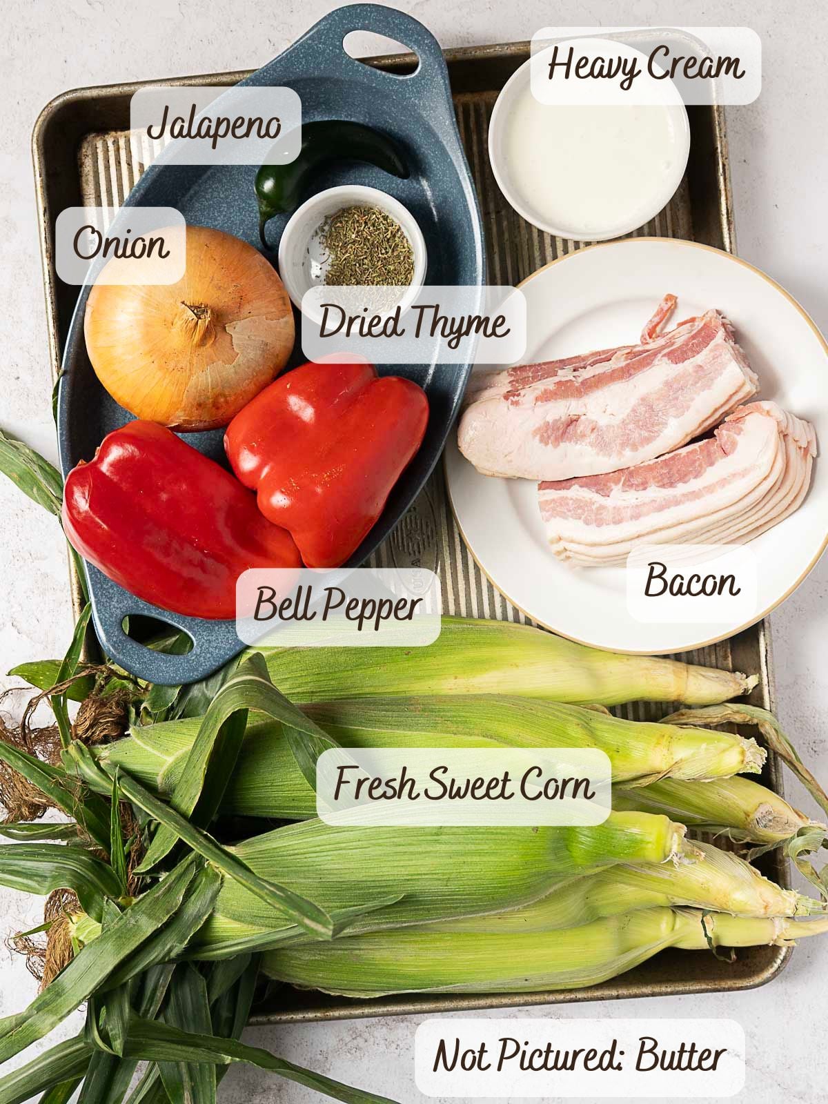 Tray with fresh sweet corn, red bell peppers, onion, jalapeno, bacon, heavy cream, dried thyme—which are all classic Maque Choux ingredients—and a note indicating butter is not pictured.