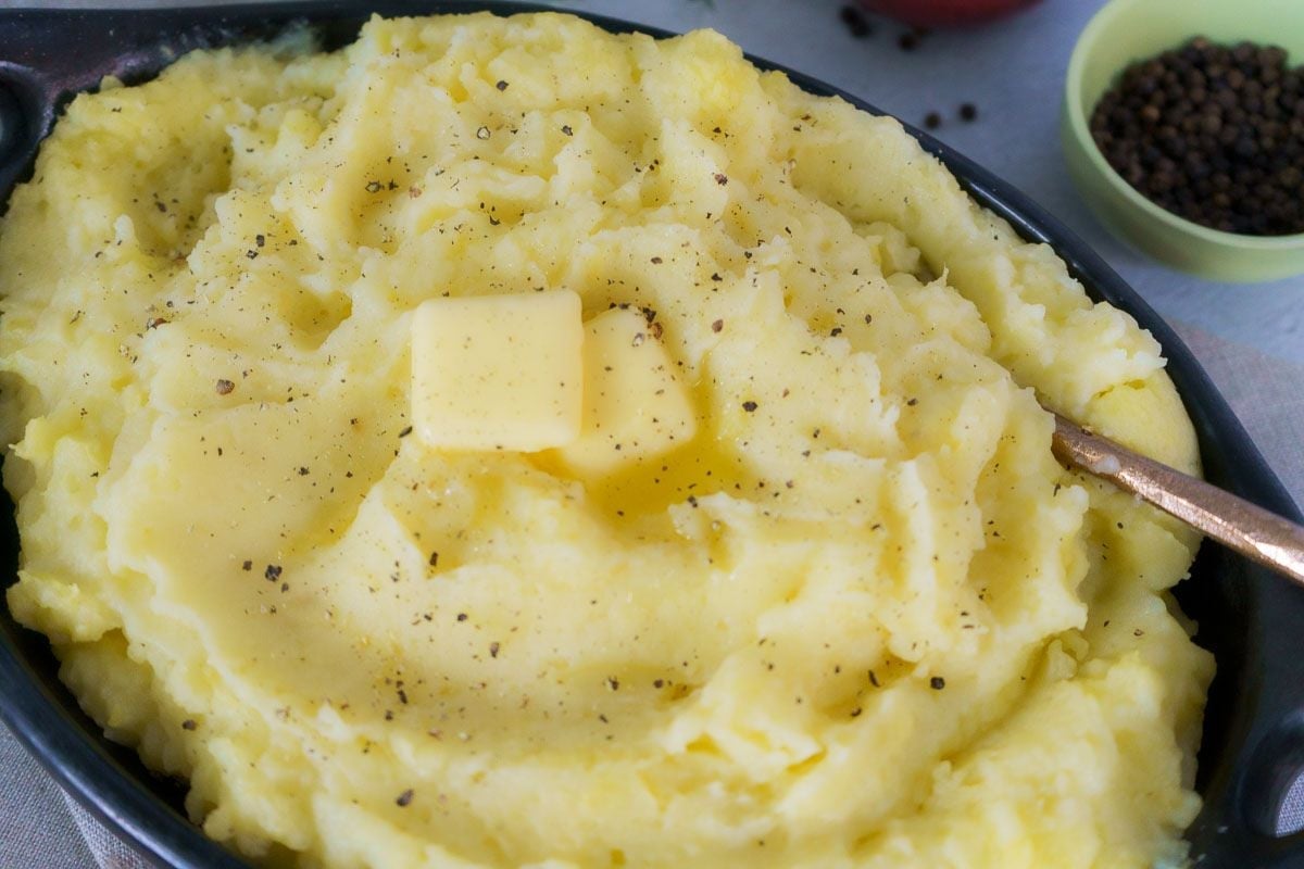 A bowl of mashed potatoes topped with a pat of butter and sprinkled with black pepper, with a spoon on the side—perfect as one of your go-to potato side dishes for any cozy fall dinner.