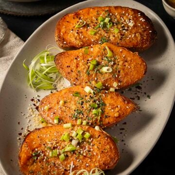 Four baked potato halves topped with chopped green onions, sesame seeds, and spices are arranged on an oval plate with a garnish of sliced green onions on the side.